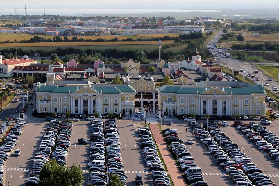 Designer Outlet Parndorf von oben: Gebäude mit grünen Dächern, dahinter die Gassen des Outlets, hinten der Neusiedler See